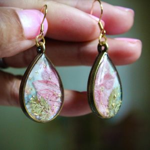 Boucles d'oreilles en forme de gouttes dorées, avec une fleur rose séchée, des fougères miniatures et des éclats dorés sur un fond pastel doux.