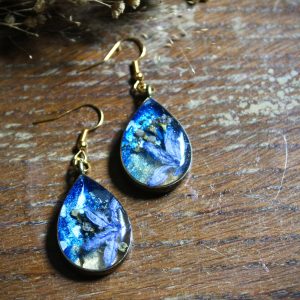 Paire de boucles d'oreilles dorées en forme de gouttes, contenant une fleur violette séchée sur un fond évoquant un paysage de plage