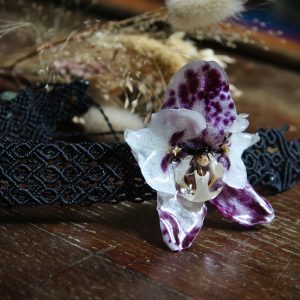 Gros plan sur un ras de cou en micro-macramé noir porté autour du cou d'une femme, orné d'une véritable fleur d'orchidée séchée aux pétales violet moucheté de blanc.
