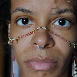 Chaîne faciale rituelle réalisée à la main, composée de perles rouges, blanches et dorées, agrémentée de petites clochettes en laiton. Elle est portée autour du visage, depuis chaque oreille jusqu'à l'arête du nez, créant un ornement à la fois spirituel et expressif.