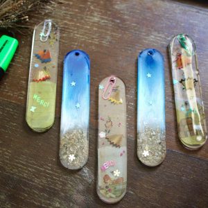 Cinq marque-pages artisanaux en résine faits main, décorés avec des copeaux de crayons, paillettes et petits mots comme "merci".