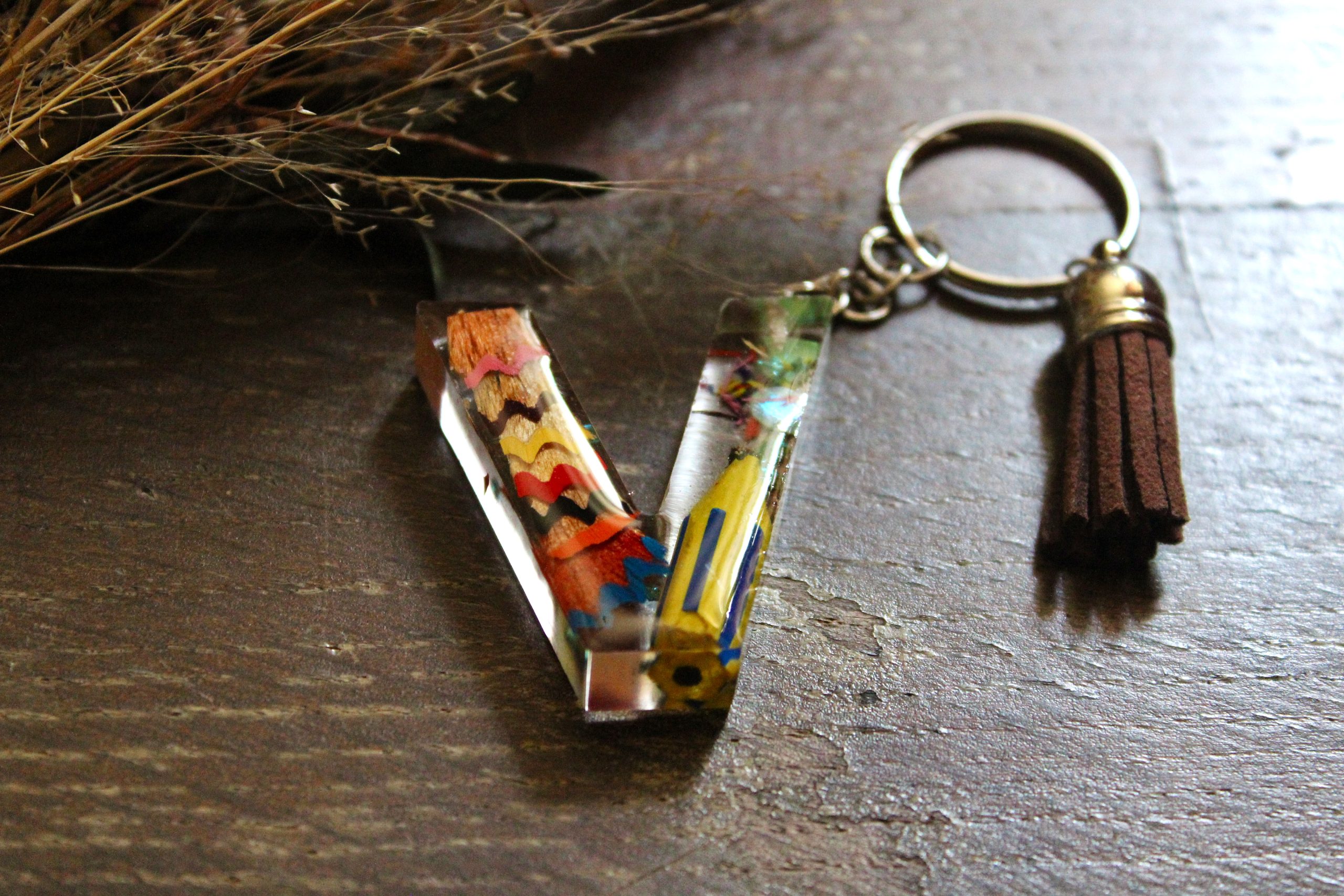 Porte-clés artisanal en résine en forme de lettre V, décoré de copeaux de crayons colorés et d'un pompon en suédine marron.