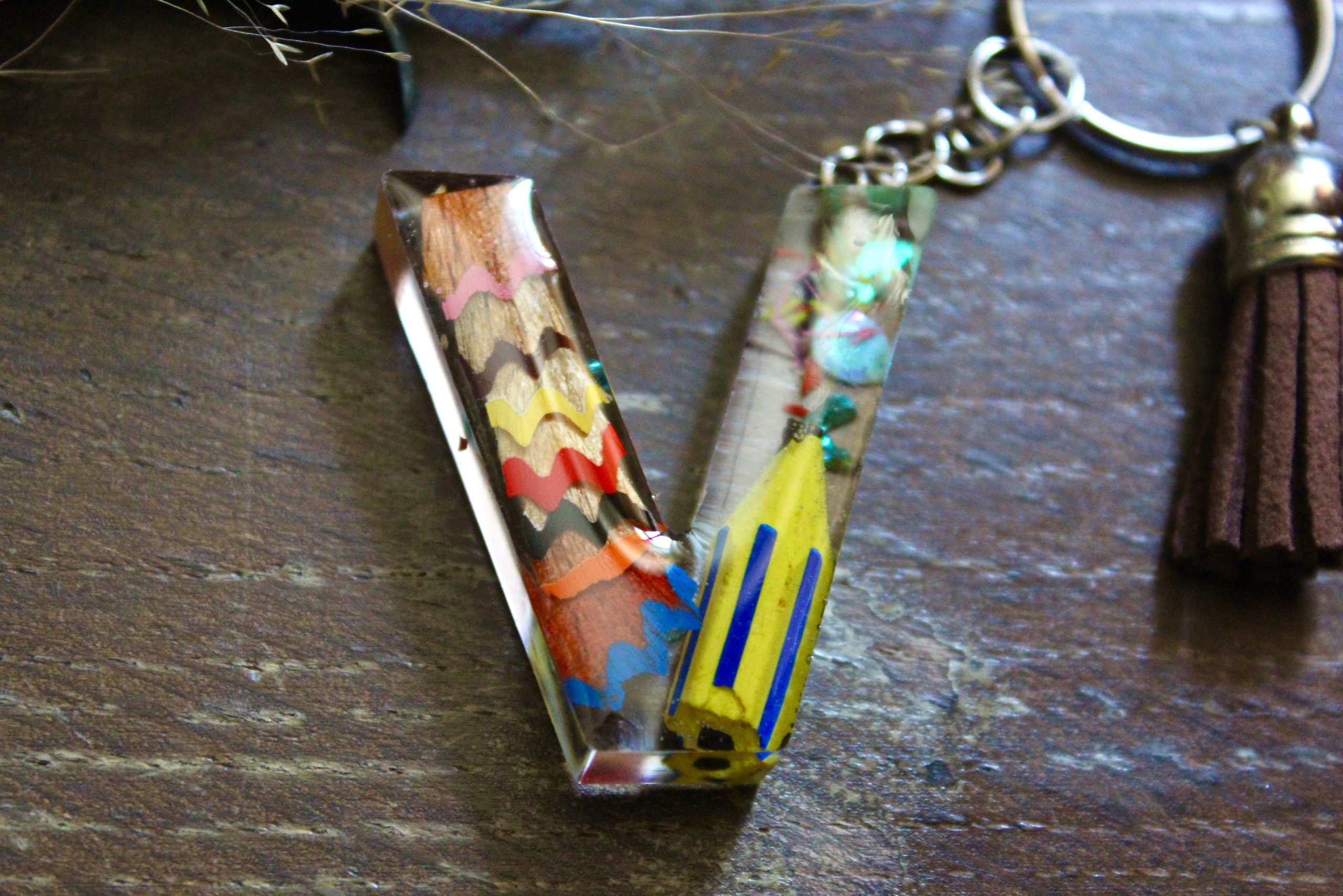 Porte-clés artisanal en résine en forme de lettre V, décoré de copeaux de crayons colorés et d'un pompon en suédine marron.