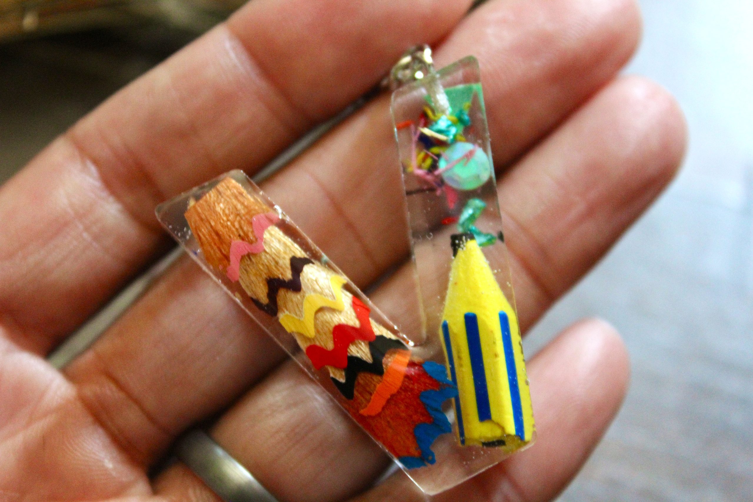 Porte-clés artisanal en résine en forme de lettre V, décoré de copeaux de crayons colorés et d'un pompon en suédine marron.