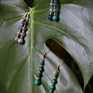 Trois paires de boucles d'oreilles pendantes en apatite, labradorite et malachite posées sur une feuille verte, version crochets dores et version crochets argentés